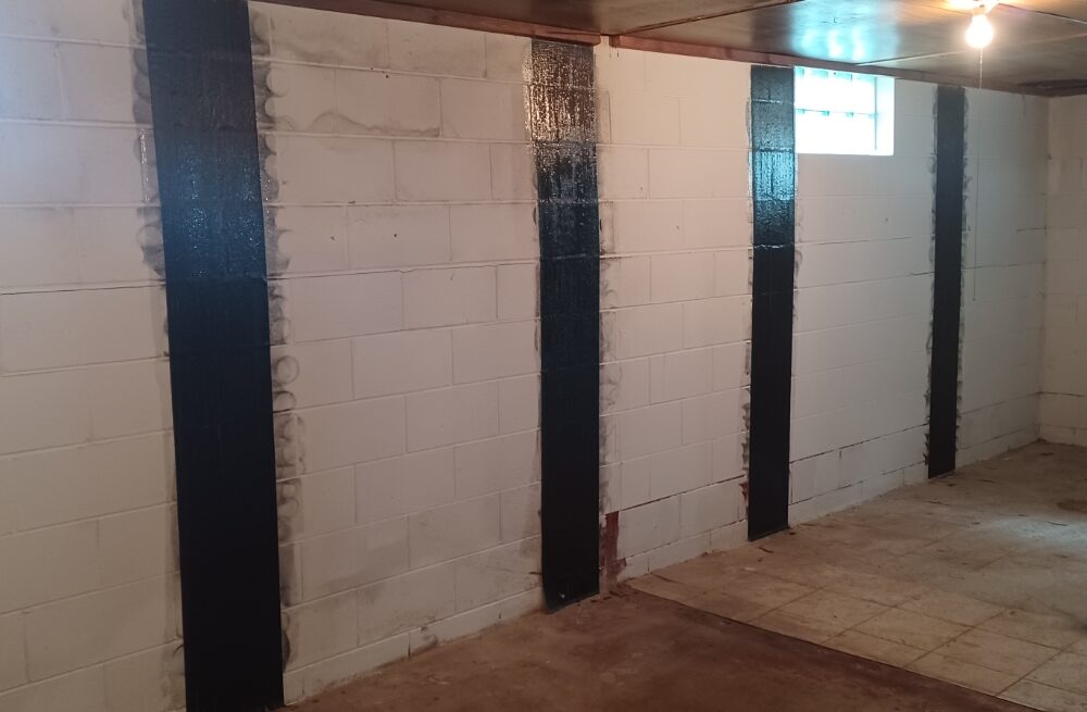 Basement room with white concrete block walls, reinforced by vertical black stripes. Two small windows let in light, creating a stark, industrial feel.